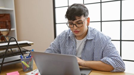 Téléchargez les photos : Jeune homme avec des lunettes de travail attentif sur ordinateur portable dans un intérieur de bureau moderne. - en image libre de droit