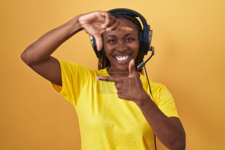 Téléchargez les photos : Femme afro-américaine écoutant de la musique à l'aide d'écouteurs souriant faisant cadre avec les mains et les doigts avec le visage heureux. concept de créativité et photographie. - en image libre de droit