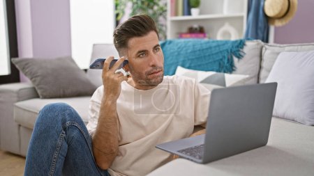 Photo for Focused young hispanic man using laptop while sitting on floor at home, intently listening to voice message on smartphone - Royalty Free Image