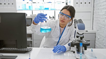 Foto de Científica hispana examinando un frasco con líquido azul en un moderno laboratorio. - Imagen libre de derechos