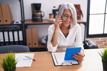 Téléchargez les photos : Femme d'affaires aux cheveux gris d'âge moyen parlant sur smartphone à l'aide d'un pavé tactile au bureau - en image libre de droit