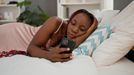 Photo for Beautiful african american woman at home, comfortably relaxing on bed in pajamas, typing message using smartphone, amidst a serene bedroom atmosphere - Royalty Free Image