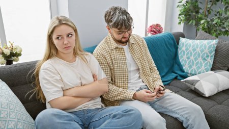 Una mujer disgustada y un hombre distraído se sientan juntos pero emocionalmente separados en un sofá en una sala de estar moderna, mostrando desafíos de relación.