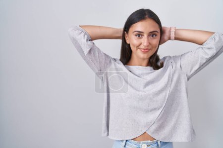Foto de Young hispanic woman standing over white background relaxing and stretching, arms and hands behind head and neck smiling happy - Imagen libre de derechos