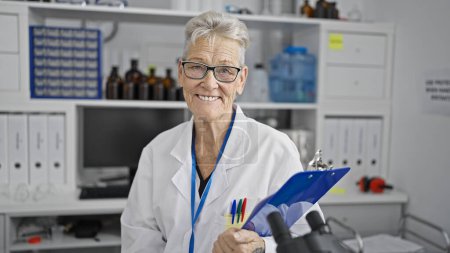 Téléchargez les photos : Femme scientifique chevronnée aux cheveux gris souriant avec confiance à un laboratoire, tout en tenant une liste de contrôle pour la recherche médicale. - en image libre de droit