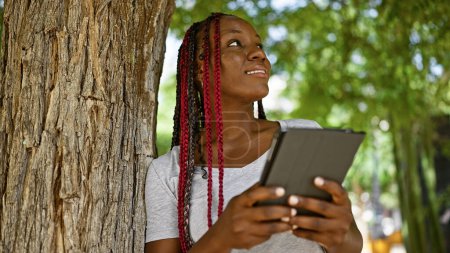 Téléchargez les photos : Femme afro-américaine confiante, élégant dans le parc de la ville, rayonne de positivité tout en utilisant le pavé tactile, son sourire prête un air de gaieté infectieuse, entouré par le vert luxuriant à l'extérieur. - en image libre de droit