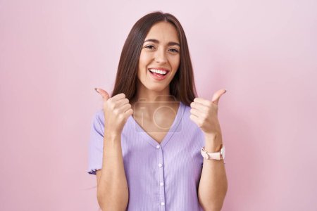Foto de Mujer hispana joven con el pelo largo de pie sobre el signo de éxito de fondo rosa haciendo gesto positivo con la mano, pulgares hacia arriba sonriendo y feliz. expresión alegre y gesto ganador. - Imagen libre de derechos