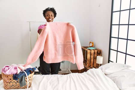 Téléchargez les photos : Femme afro-américaine tenant un pull faisant la lessive dans la chambre - en image libre de droit