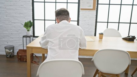 Photo for Relaxed yet successful, a young hispanic man redefines business norms, sitting backwards on a table, engrossed in work at his indoor office. a refreshing take on a professional portrait. - Royalty Free Image