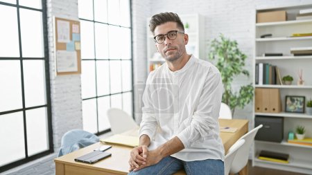Photo for Young hispanic man in business attire working undisturbed, caught in a relaxed yet serious posture facing the camera amidst the indoor panorama of his office. - Royalty Free Image