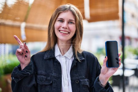 Foto de Sonriente, joven rubia apuntando hacia un lado con una mano, felizmente sosteniendo su teléfono inteligente mientras está de pie en la terraza de una cafetería. - Imagen libre de derechos