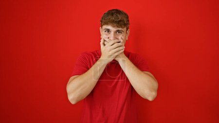 Photo for Cheerful young hispanic guy confidently covering mouth with hands over a vibrant isolated red background, exuding confidence and happiness - Royalty Free Image