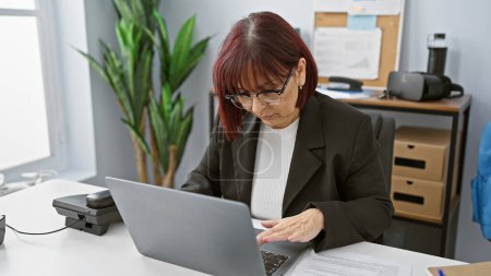 Téléchargez les photos : Femme hispanique mature travaillant concentrée dans un cadre de bureau moderne - en image libre de droit