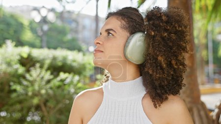 Téléchargez les photos : Femme hispanique avec des cheveux bouclés portant des écouteurs aime la musique dans un parc ensoleillé en plein air - en image libre de droit