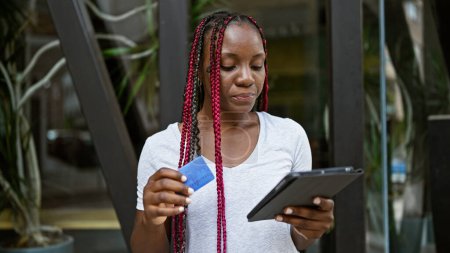 Téléchargez les photos : Belle femme afro-américaine avec des tresses faisant des achats décontractés sur son pavé tactile dans une rue ensoleillée de la ville, effectuant des paiements en ligne avec carte de crédit - en image libre de droit