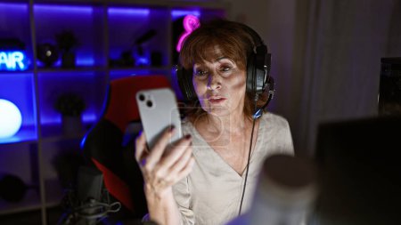 Photo for A mature woman with headphones uses a smartphone in a neon-lit gaming room at night, portraying modern senior leisure activities. - Royalty Free Image