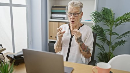 Photo for Serene grey-haired senior woman, an elegant business worker, crossing her fingers for luck while working on her laptop at her desk in the office. - Royalty Free Image