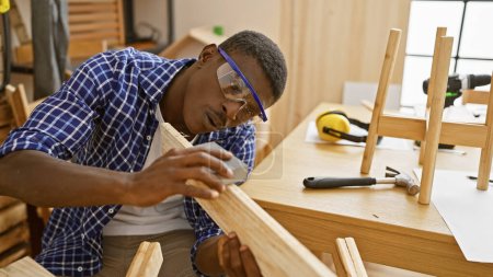 Foto de Hombre negro guapo lijando madera diligentemente en un taller de carpintería bien equipado. - Imagen libre de derechos