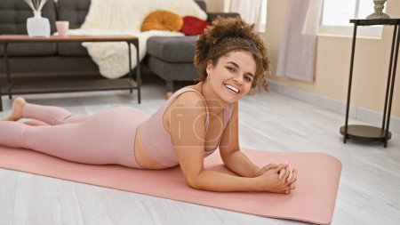 Foto de Mujer hispana joven con el pelo rizado sonriendo mientras está acostada en una esterilla de yoga rosa en su acogedora sala de estar. - Imagen libre de derechos