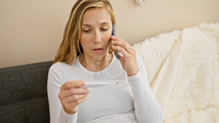 Foto de Una joven mujer caucásica preocupada hablando por teléfono mientras comprueba su temperatura en casa, creando una escena de enfermedad potencial. - Imagen libre de derechos