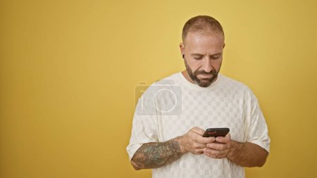 Foto de Joven guapo, absorto en una conversación seria de texto, usando tecnología de teléfonos inteligentes, aislado sobre un fondo amarillo vibrante. - Imagen libre de derechos
