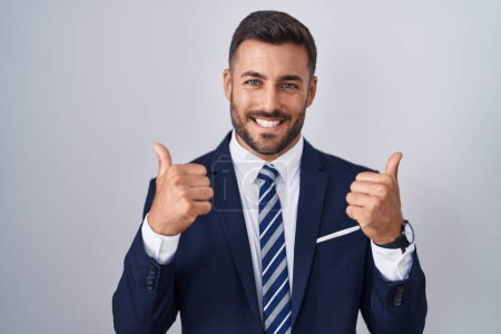 Foto de Hombre hispano guapo vistiendo traje y corbata signo de éxito haciendo gesto positivo con la mano, pulgares arriba sonriendo y feliz. expresión alegre y gesto ganador. - Imagen libre de derechos