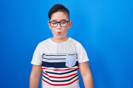 Foto de Joven niño hispano de pie sobre fondo azul haciendo cara de pez con los labios, loco y cómico gesto. expresión divertida. - Imagen libre de derechos