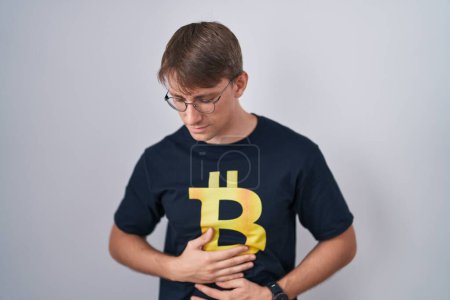 Photo for Caucasian blond man wearing bitcoin t shirt with hand on stomach because indigestion, painful illness feeling unwell. ache concept. - Royalty Free Image