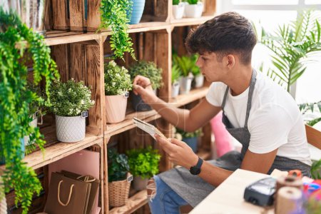 Téléchargez les photos : Jeune hispanique adolescent fleuriste lecture carnet toucher plante à la boutique de fleurs - en image libre de droit