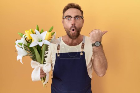 Photo for Middle age man with beard florist shop holding flowers surprised pointing with hand finger to the side, open mouth amazed expression. - Royalty Free Image