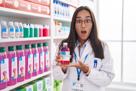 Téléchargez les photos : Jeune femme hispanique travaillant à la pharmacie pharmacie tenant du sirop en état de choc, à l'air sceptique et sarcastique, surprise avec la bouche ouverte - en image libre de droit