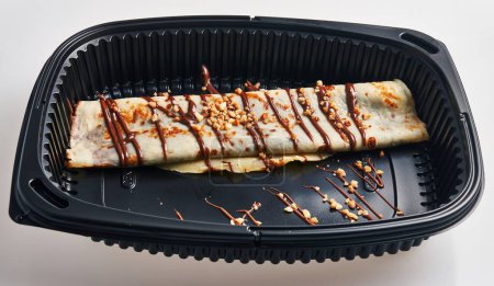 Photo for A delicious crepe with chocolate and nuts in a black takeout container invites indulgence. - Royalty Free Image