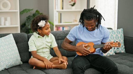 Foto de Padre e hija afroamericanos sentados en sofá enseñando a tocar ukelele en casa - Imagen libre de derechos
