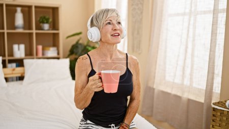 Photo for A smiling middle-aged woman with grey hair listening to music on headphones while holding a mug in a bedroom setting. - Royalty Free Image