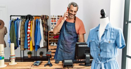 Foto de Asistente de tienda hombre de pelo gris de mediana edad usando el ordenador hablando en el teléfono inteligente en la tienda de ropa - Imagen libre de derechos