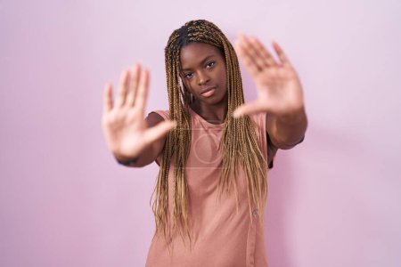 Téléchargez les photos : Femme afro-américaine avec des cheveux tressés debout sur fond rose faisant cadre en utilisant les mains paumes et les doigts, perspective de la caméra - en image libre de droit