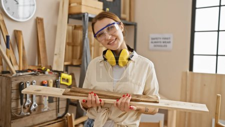 Foto de Mujer sonriente con gafas de seguridad sosteniendo madera en un taller de carpintería bien equipado - Imagen libre de derechos