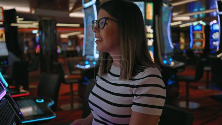 Photo for A smiling young woman in glasses plays slot machines in the vibrant ambiance of a casino. - Royalty Free Image