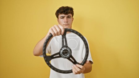 Photo for Handsome young hispanic teenager with a serious expression mastering the steering wheel as a relaxed driver, against an isolated yellow background. - Royalty Free Image
