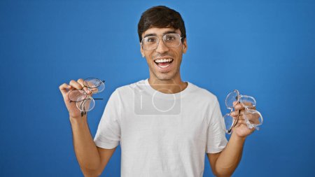 Téléchargez les photos : Joyeux jeune hispanique gars, clignotant avec confiance un sourire charmant tout en tenant des lunettes, debout isolé sur un fond bleu frais - en image libre de droit