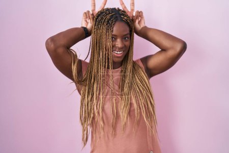 Téléchargez les photos : Femme afro-américaine avec des cheveux tressés debout sur fond rose posant drôle et fou avec les doigts sur la tête comme des oreilles de lapin, souriant gai - en image libre de droit