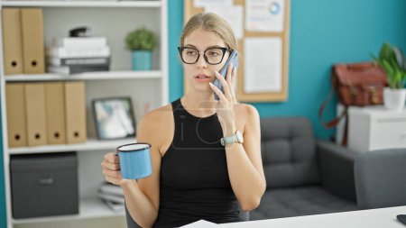 Téléchargez les photos : Jeune femme blonde femme d'affaires parlant sur smartphone boire du café au bureau - en image libre de droit