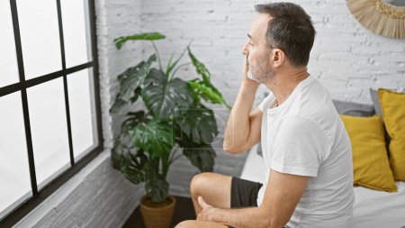 Téléchargez les photos : Homme d'âge moyen avec les cheveux gris assis sur le lit regardant jeter la fenêtre à la chambre - en image libre de droit