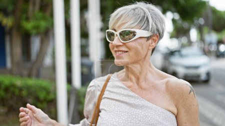 Foto de Mujer hispana madura con pelo gris y gafas de sol sonriendo en una calle urbana soleada. - Imagen libre de derechos