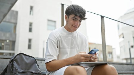 Téléchargez les photos : Jeune étudiant hispanique ensoleillé, envoyant joyeusement des SMS sur son smartphone assis sur les escaliers du campus de la ville, rayonnant de confiance avec son sourire magnétique - en image libre de droit