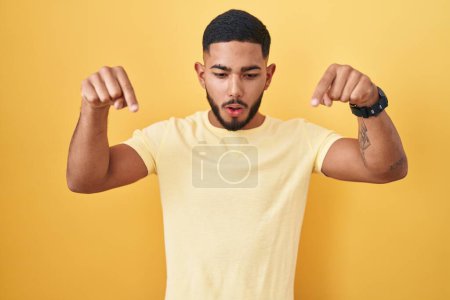 Téléchargez les photos : Young hispanic man standing over yellow background pointing down with fingers showing advertisement, surprised face and open mouth - en image libre de droit
