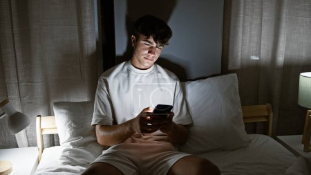 Téléchargez les photos : Jeune adolescent hispanique utilisant smartphone assis sur le lit dans la chambre - en image libre de droit