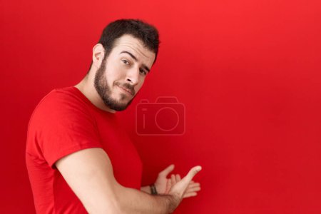 Téléchargez les photos : Jeune homme hispanique portant un t-shirt rouge décontracté invitant à entrer souriant naturel à la main ouverte - en image libre de droit