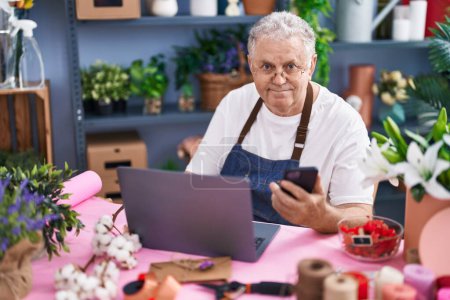 Foto de Floristería hombre de pelo gris de mediana edad usando teléfono inteligente y portátil en floristería - Imagen libre de derechos