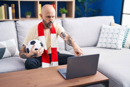 Photo for Hispanic man with tattoos watching football match hooligan holding ball on the laptop pointing with finger to the camera and to you, confident gesture looking serious - Royalty Free Image